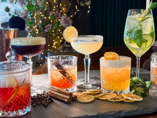 Hurley House - A selection of colorful cocktails garnished with citrus slices, mint, and cinnamon sticks, displayed on a slate tray with dried fruit and fairy lights in the background.