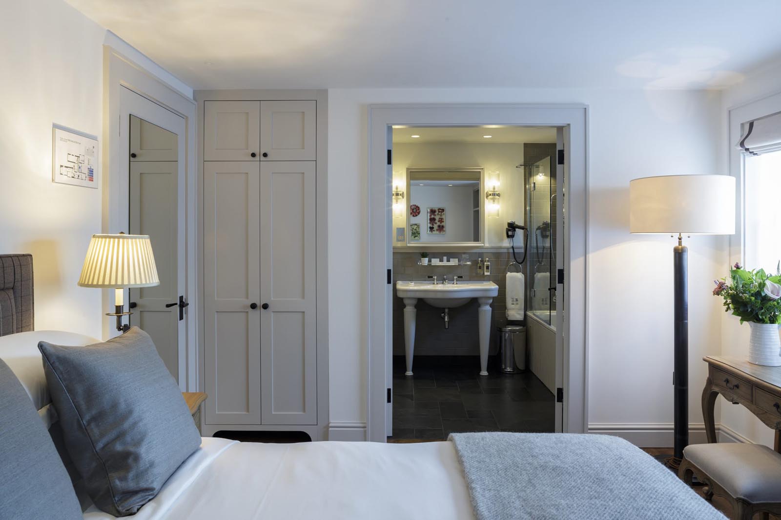 Hurley House - A cozy bedroom with a neatly made bed, gray throw blanket, and light gray pillows. A bedside table with a lamp is beside it. Across is an open bathroom with white fixtures. A tall floor lamp and a bouquet of flowers are near the window.