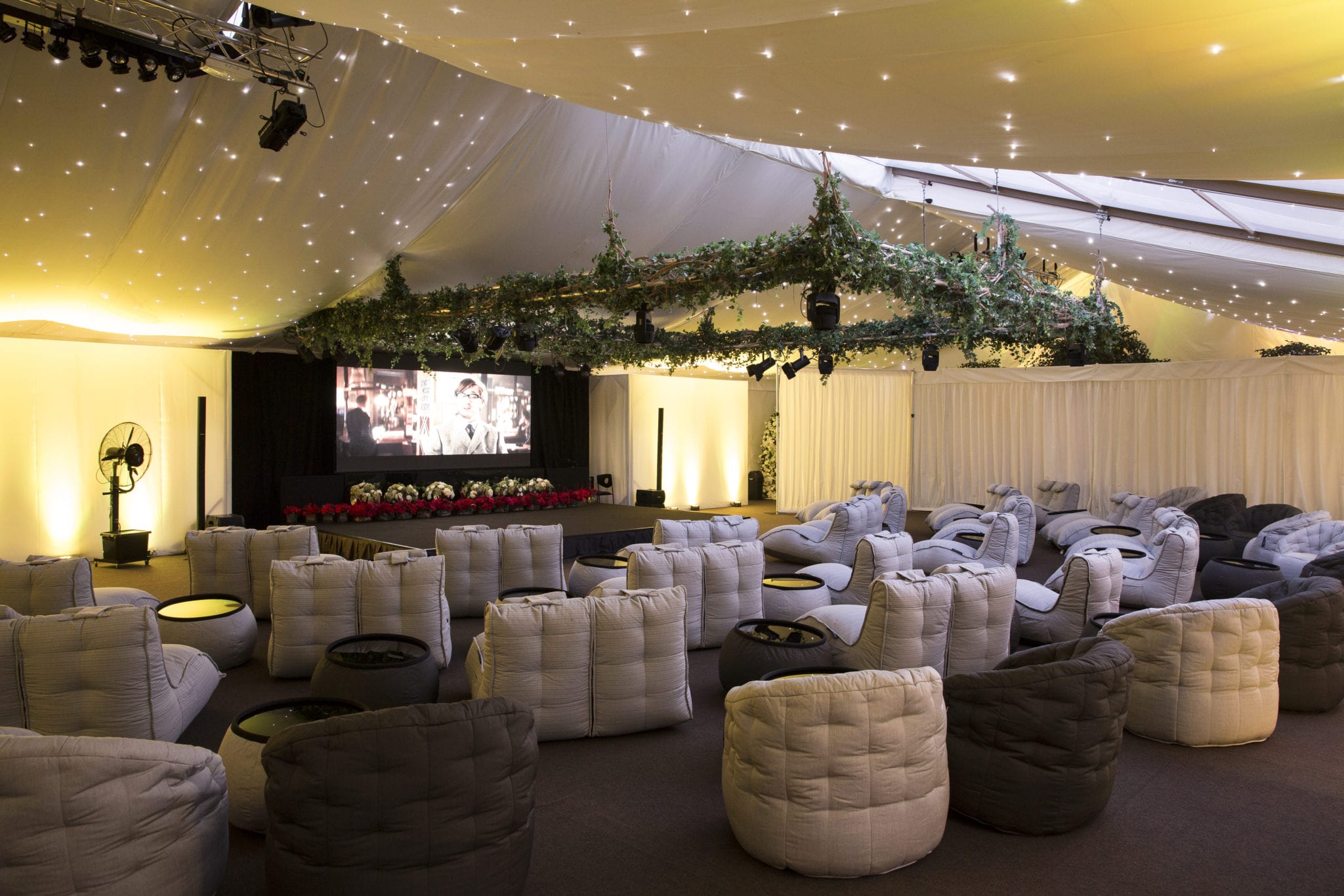 Hurley House - A large event tent with a plush seating area, featuring gray and beige bean bags arranged in rows facing a stage with a screen. The ceiling is decorated with hanging greenery and soft, sparkling lights, creating a cozy ambiance.