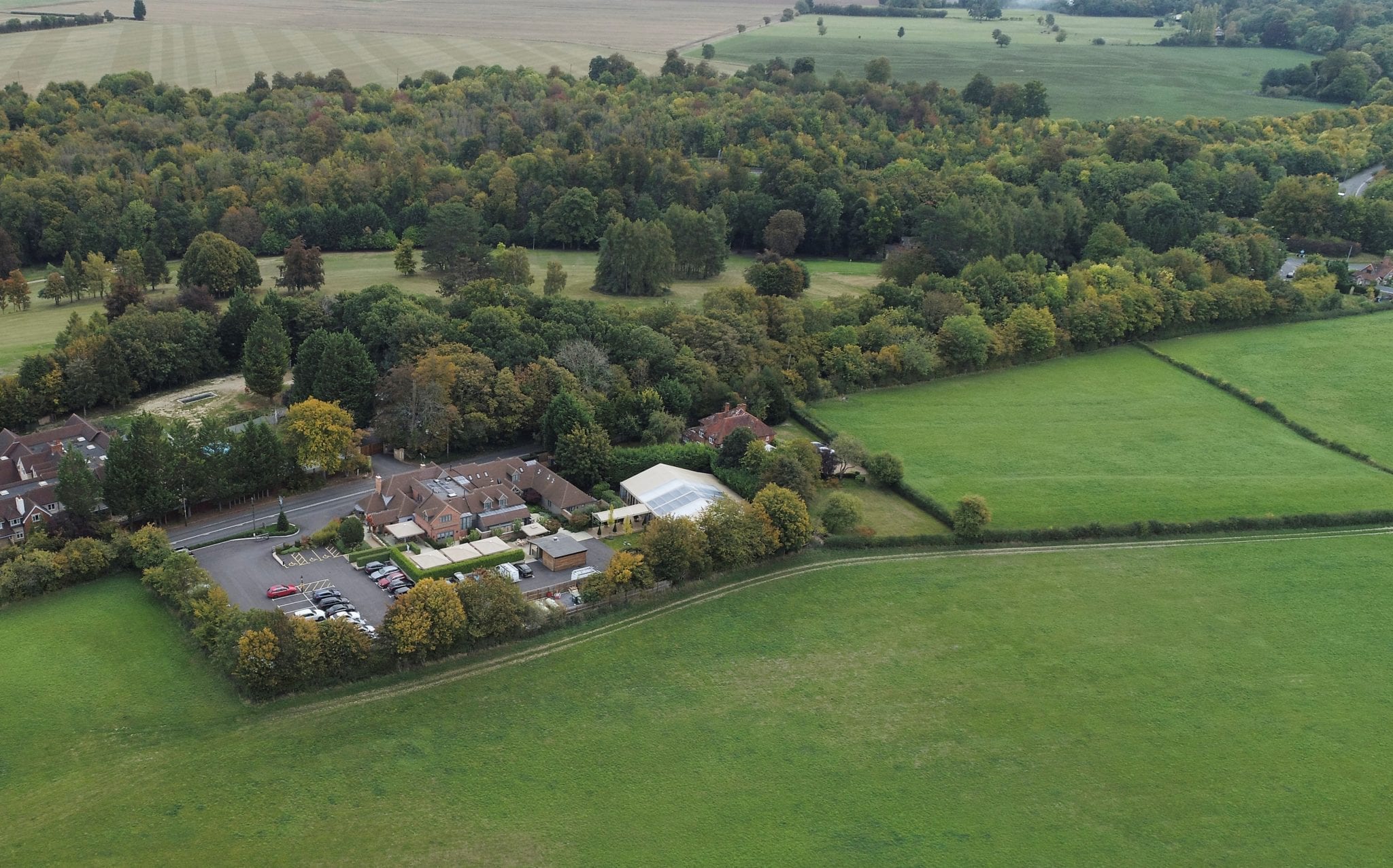 Hurley House - Aerial view of a rural landscape with a cluster of buildings surrounded by trees and open land. The area features fields and dense woodland, with a small parking lot adjacent to the buildings. Hills and more trees are visible in the background.