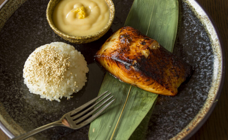 Hurley House - A plate with grilled fish on a green leaf, a serving of white rice topped with sesame seeds, a small bowl of creamy sauce, and a fork, all set on a dark ceramic plate on a wooden table.
