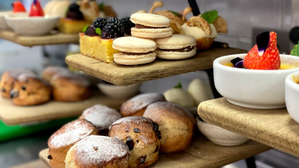 Hurley House - A close-up of wooden trays with assorted pastries, including scones dusted with powdered sugar, macarons, sponge cake with berries, and desserts garnished with fresh fruit and mint.