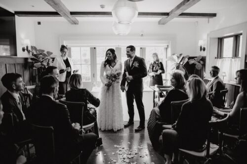 Hurley House - A black and white photo of a wedding ceremony. A bride and groom stand together, smiling, at the front of a room filled with seated guests. Natural light streams in from windows behind them, illuminating rose petals scattered on the floor.