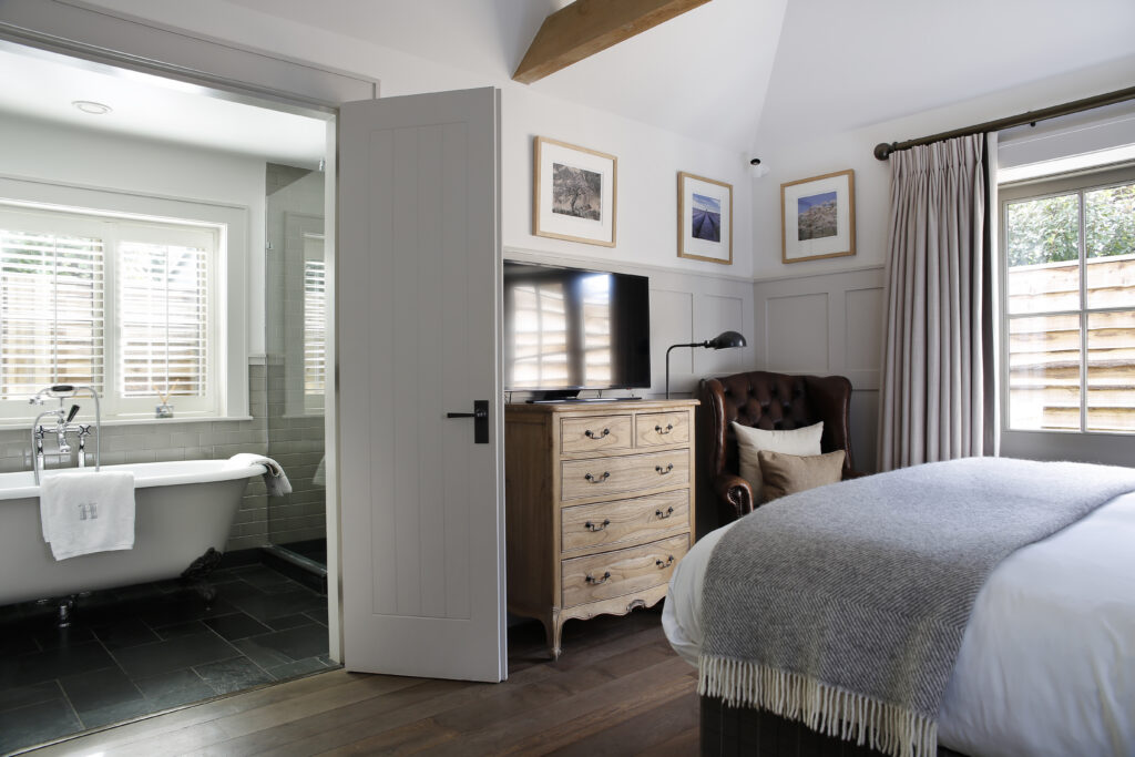 Hurley House - A cozy bedroom at this luxury wedding venue in Berkshire features a neatly made bed, gray blanket, wooden dresser with TV, and an armchair. The en suite bathroom has a freestanding bathtub, white shutters, gray tile flooring, and framed photos on the walls.