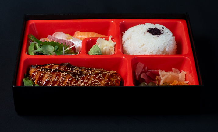 Hurley House - A Japanese bento box with white rice topped with seasoning, teriyaki-glazed salmon, mixed sashimi, pickled vegetables, and a green salad, all arranged in separate red compartments.