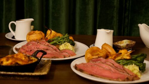 Hurley House - Plates of sliced roast beef with Yorkshire pudding, broccoli, potatoes, and gravy served on a wooden table, with sauce jugs and side dishes, in front of a dark green curtain.