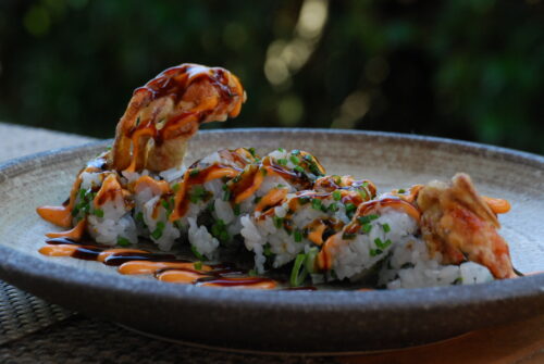 Hurley House - A close-up of a sushi roll topped with sauce and garnished with herbs, featuring a piece of fried seafood, served on a rustic ceramic plate with a blurred green background.