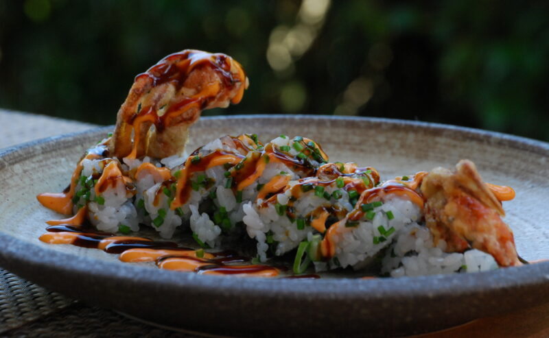 Hurley House - A close-up of a sushi roll topped with sauce and garnished with herbs, featuring a piece of fried seafood, served on a rustic ceramic plate with a blurred green background.