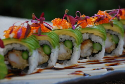Hurley House - A close-up of sushi rolls topped with avocado slices, orange fish roe, and microgreens, with asparagus and tempura filling, arranged on a plate drizzled with dark and light sauces.
