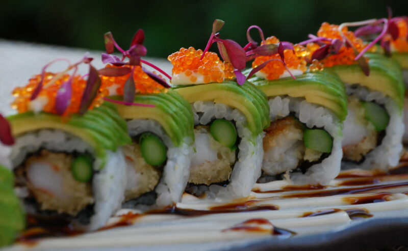 Hurley House - A close-up of sushi rolls topped with avocado slices, orange fish roe, and microgreens, with asparagus and tempura filling, arranged on a plate drizzled with dark and light sauces.
