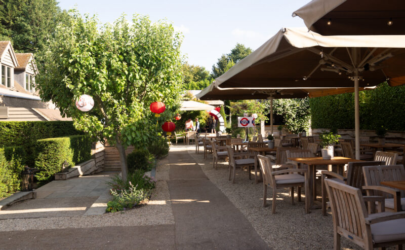Hurley House - Outdoor restaurant seating area with wooden tables and chairs under large umbrellas, surrounded by greenery and trees decorated with red lanterns on a sunny day. Pathway runs through the middle of the area.