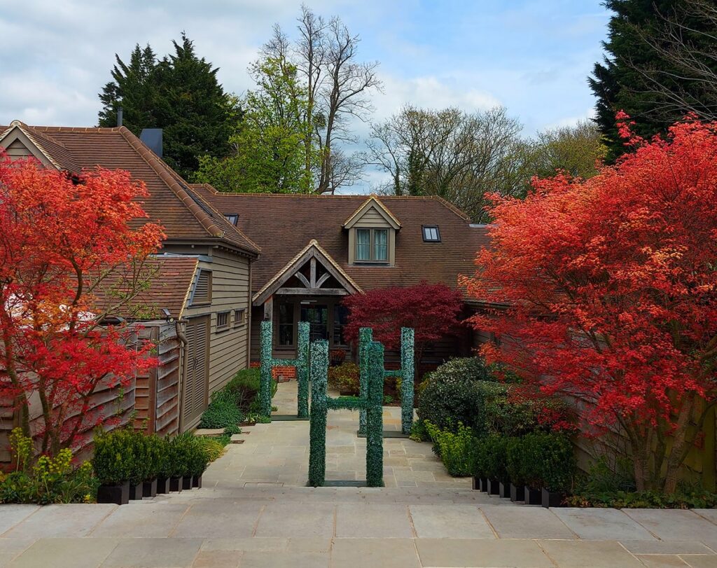 Hurley House - A house with steep gabled roofs is framed by vibrant red-orange trees and green shrubs along a paved pathway under a partly cloudy sky.
