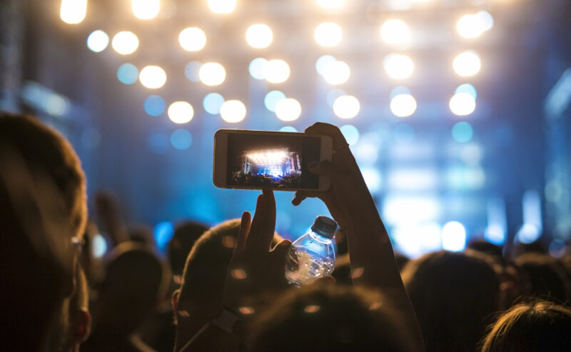Hurley House - A person in a crowd records a brightly lit concert stage at Whats on at Hurley House with a smartphone, holding a water bottle. The out-of-focus image highlights stage lights and captures the lively concert atmosphere.
