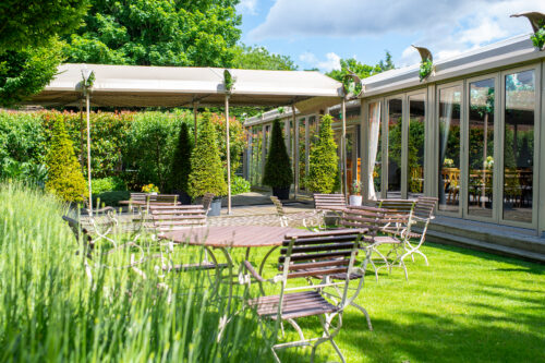 Hurley House - Outdoor seating area with metal tables and chairs on a green lawn, shaded by a canopy and surrounded by trimmed bushes and trees next to a building with large glass windows on a sunny day.