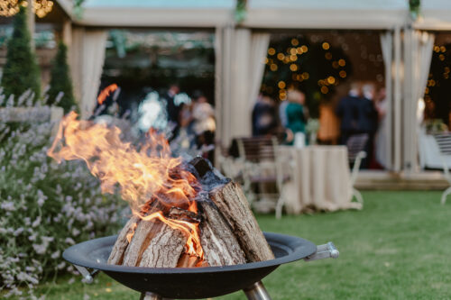 Hurley House - A close-up of a fire pit with burning logs in a grassy outdoor setting, with blurred people, tables, and string lights in the background, suggesting a festive event or gathering.