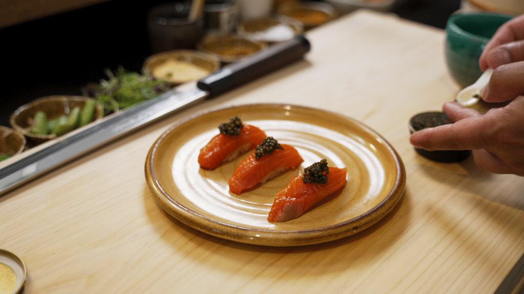 Hurley House - Three pieces of salmon sushi topped with black caviar are neatly arranged on a round ceramic plate. A hand is seen on the right, spooning more caviar from a small bowl. The scene is set on a wooden counter.
