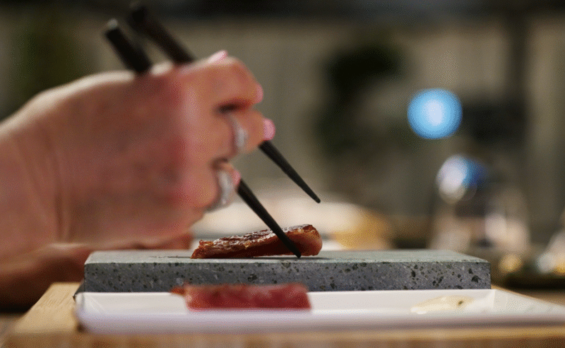 Hurley House - A close-up of a hand holding chopsticks, picking up a piece of sushi from a stone plate. The background is blurred, focusing on the sushi and chopsticks.