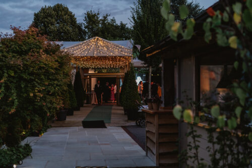 Hurley House - A warmly lit event tent with string lights is nestled among greenery at dusk. People are visible inside the tent, and the scene has a cozy, inviting atmosphere with trees and plants framing the walkway.