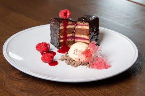 Hurley House - A plated dessert featuring layered chocolate-covered cake with fruit filling, garnished with raspberries, a scoop of vanilla ice cream, chocolate crumbles, and pink sugar tuile on a white plate.