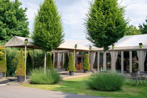 Hurley House - A garden venue with tall manicured trees, large windows, and a covered walkway leading to an entrance. Outdoor seating and lush greenery surround the building under a partly cloudy sky.
