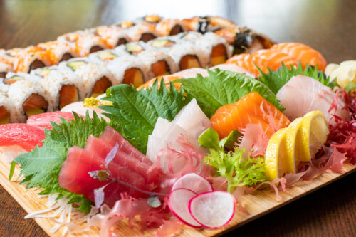 Hurley House - A wooden platter featuring assorted sashimi slices including tuna, salmon, and yellowtail, garnished with lemon, greens, and radishes, with sushi rolls in the background.