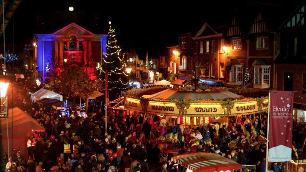 Hurley House - A festive Christmas market at night with a large crowd, decorated stalls, a carousel, a lit Christmas tree, and buildings illuminated in colorful lights. A banner reads “Henley Christmas Festival.”.