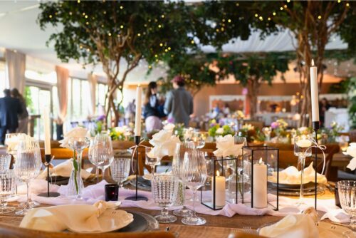 Hurley House - A beautifully decorated banquet table set for an event, featuring elegant glassware, white roses, candles, and plates, with softly lit trees and blurred guests in the background.