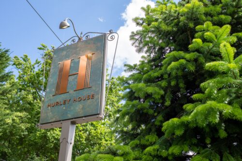 Hurley House - A large sign reading “Hurley House” with a big copper letter H stands among lush green trees under a partly cloudy sky.