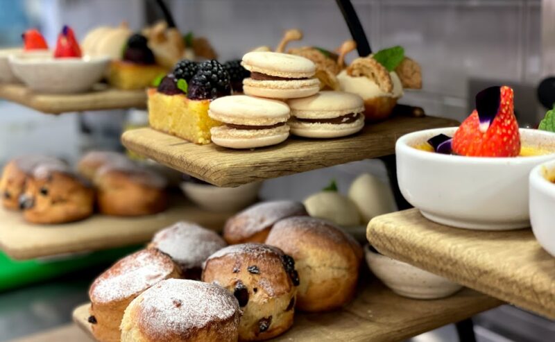 Hurley House - A variety of pastries, including scones, macarons, cakes, and fruit-topped desserts, are artfully arranged on wooden display stands in a bakery or café setting.