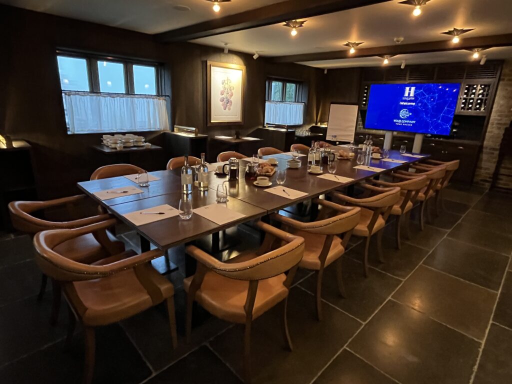 Hurley House - A conference room at a Premier Meeting & Conference Venue in Berkshire features a long wooden table set for a meeting, surrounded by leather chairs, water bottles, notepads, and a large screen. Warm lighting creates a professional yet cozy atmosphere.