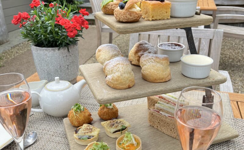 Hurley House - A three-tiered tray with assorted pastries, scones, sandwiches, and desserts is set on an outdoor table with rosé wine, a teapot, flowers, and greenery in the background.