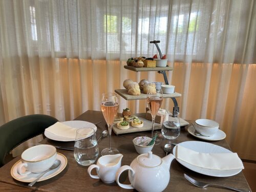 Hurley House - A table set for afternoon tea with two place settings, a white teapot, glasses of pink sparkling wine, and a three-tiered stand with assorted pastries, scones, and sandwiches. Light filters through sheer curtains behind.