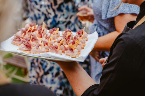 Hurley House - A person holds a white tray with neatly arranged appetizers topped with meat and garnish. People in patterned clothing are seen in the blurred background, suggesting a social or outdoor event.