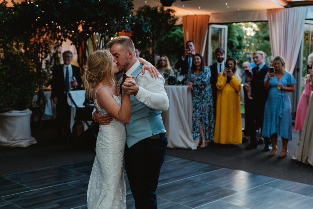 Hurley House - At a luxury wedding venue in Berkshire, a bride and groom share their first dance. Guests stand around watching, some taking photos. The elegantly decorated space, adorned with trees and draped curtains, creates a festive atmosphere perfect for this unforgettable moment.