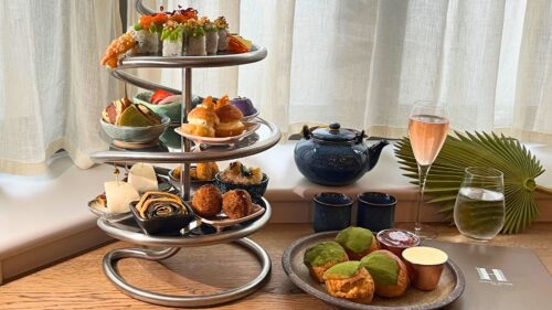 Hurley House - A modern spiral stand holds assorted sushi, savory snacks, and desserts beside a plate of matcha scones, a teapot, a glass of sparkling drink, and a glass of water on a wooden table by a window with sheer curtains.
