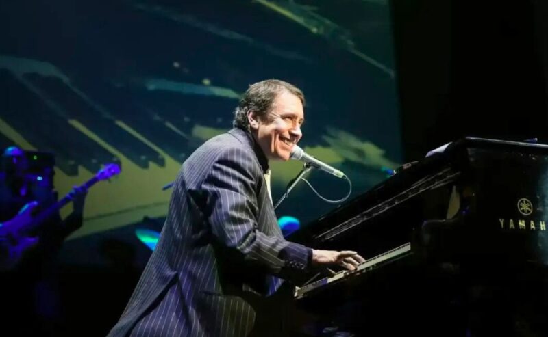 Hurley House - A man in a striped suit smiles while playing a Yamaha grand piano and singing into a microphone on stage, with musicians and a large piano graphic in the background—capturing the vibrant spirit of Whats on at Hurley House.