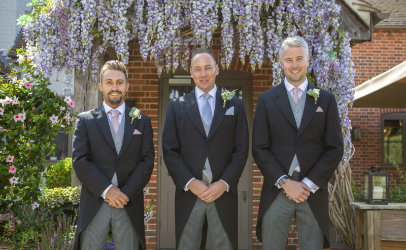Hurley House - Three men in formal suits stand smiling in front of a brick building adorned with blooming wisteria, an exquisite backdrop for a luxury wedding venue in Berkshire. They each wear matching dark jackets, gray vests, and ties, complete with boutonnieres on their lapels.