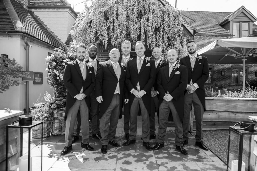 Hurley House - A group of eight men in formal suits stand together outdoors, posing for a photo at a luxury wedding venue in Berkshire. They are in front of a lush, white flowering tree on a sunny day, with brick buildings and a patio area elegantly framing the backdrop.