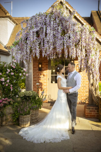 Hurley House - A bride and groom stand under a wooden arch covered with cascading purple flowers, gazing at each other affectionately outside a brick building on a sunny day. Baskets of flowers surround the couple.