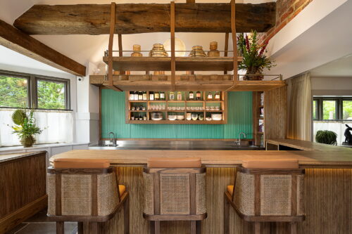 Hurley House - A rustic kitchen with wooden beams and a wood-paneled bar counter reminiscent of an omakase sushi bar, the best in Berkshire. Three rattan bar stools are lined up at the counter. Shelves above are stocked with jars and bottles. A green backsplash and flowers add color, while large windows offer natural light.