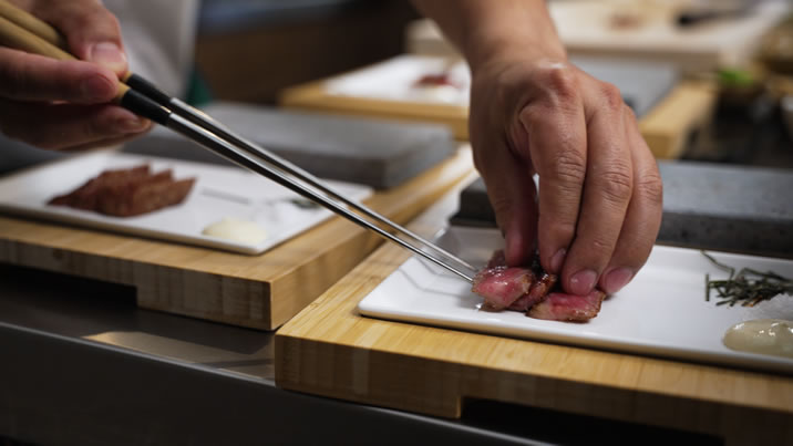 Hurley House - A chef uses tongs to carefully place slices of seared meat onto a rectangular white plate, which also has a small portion of sauce and garnish, all set on a wooden board.