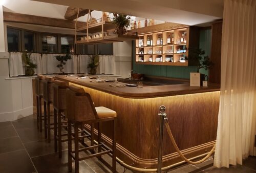 Hurley House - A cozy, dimly lit bar with a wooden counter and four high-backed chairs. Shelves with bottles and glasses are in the background, and hanging plants add a touch of greenery. A rope barrier and curtain frame the area.
