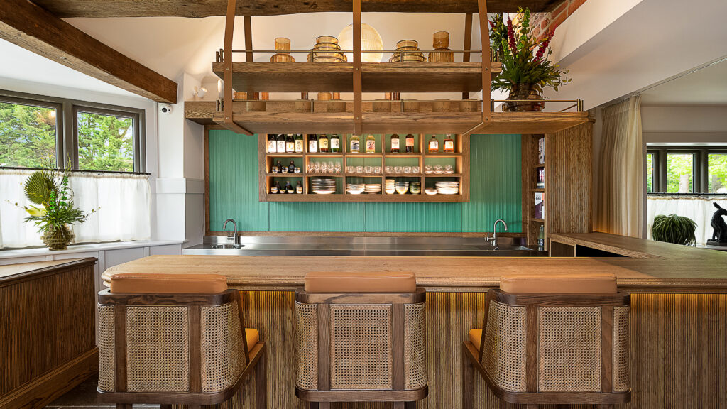 Hurley House - A stylish kitchen with three rattan bar stools, a wooden island, green paneling, open shelves with dishes and bottles, and hanging lights, surrounded by large windows and indoor plants.