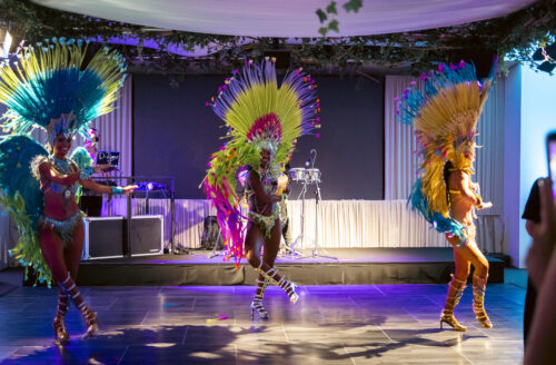 Hurley House - Three dancers in colorful feathered costumes perform energetically on a stage with vibrant lighting. The background has musical instruments and decorations with greenery above. An audience member is visible in the foreground.