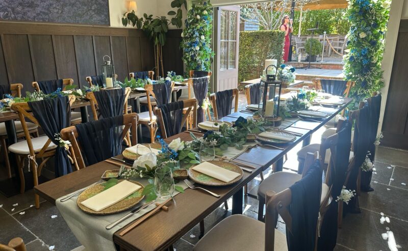 Hurley House - A luxurious wedding reception table set in a rustic Berkshire venue. Wooden chairs with dark drapes surround a table adorned with greenery, flowers, and lanterns. Light streams in from an open door, revealing an outdoor area lush with greenery.