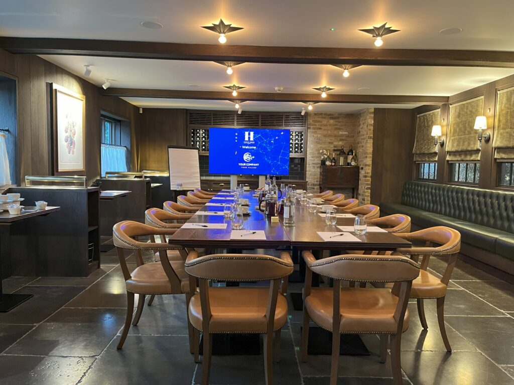 Hurley House - A well-lit conference room with a long table set for a meeting, featuring notebooks, glasses, and water bottles. A large screen at the end displays a blue presentation slide. Wooden chairs and modern decor complete the room.