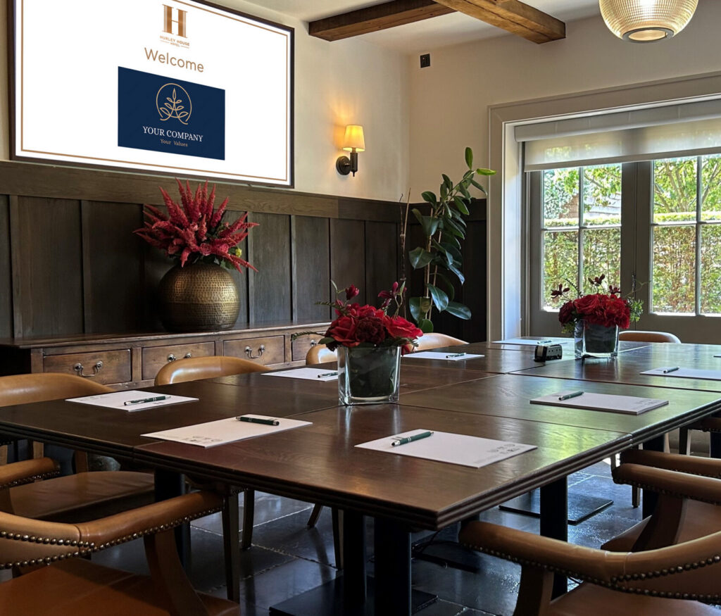 Hurley House - A well-lit conference room with a large table set for a meeting, leather chairs, notepads, pens, red flower centerpieces, and a screen displaying a welcome message near large windows with a garden view.