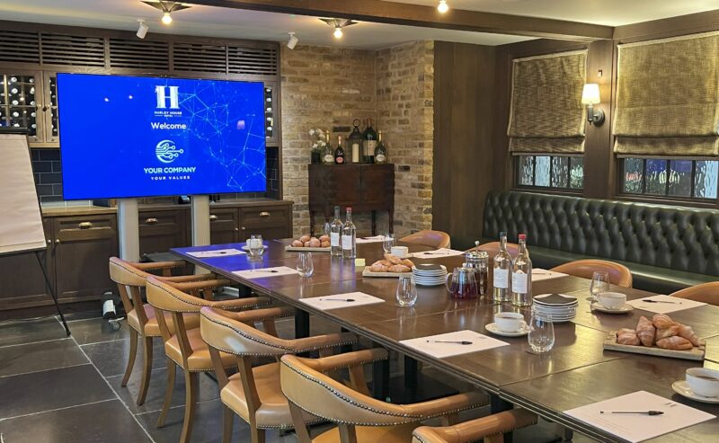 Hurley House - A modern conference room with a long table set for a meeting, surrounded by tan chairs. There are glasses, bottles, and pastries on the table. A screen displays a welcome message. The room has brick walls and soft lighting.