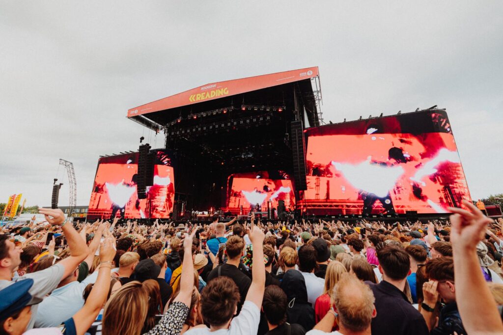 Hurley House - A large crowd faces an outdoor music stage with “Reading” on the roof. People raise their hands as bright red visuals display on big screens during a lively festival—just one example of What’s on at Hurley House this season.