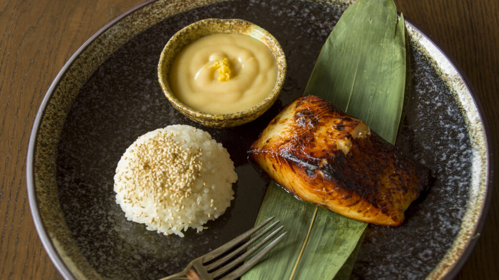Hurley House - A plate with a portion of grilled fish on a green leaf, a mound of white rice topped with sesame seeds, and a small bowl of creamy yellow sauce, all arranged on a dark ceramic plate with a fork nearby.
